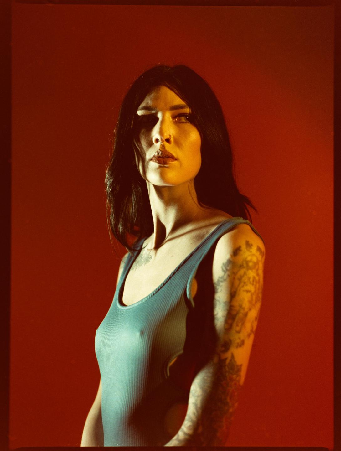 A woman with long dark hair and tattoos on her arms is looking to the side against a red background.