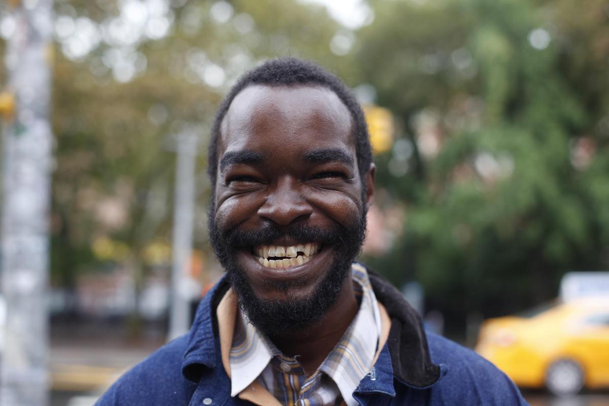 A man with a beard and a plaid shirt smiles at the camera on a city street.