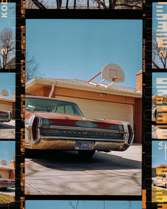 A rusty red car is parked in a driveway in front of a garage with a basketball hoop on the roof.