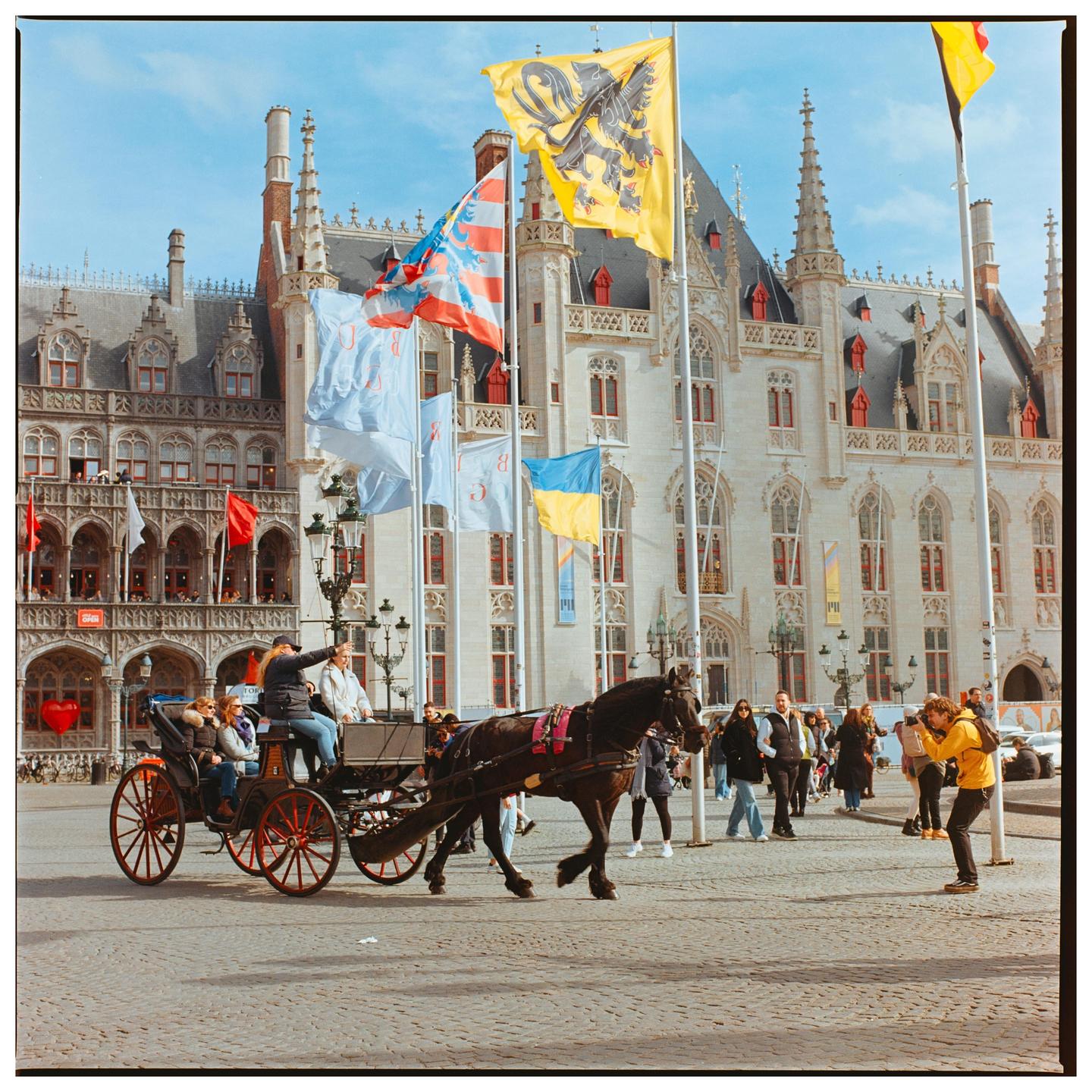 A horse-drawn carriage with people rides on a cobblestone street in front of a large ornate building with flags flying.