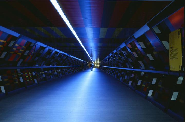 A long, narrow, blue-lit tunnel with colorful panels on the walls and ceiling has a person walking in the distance.