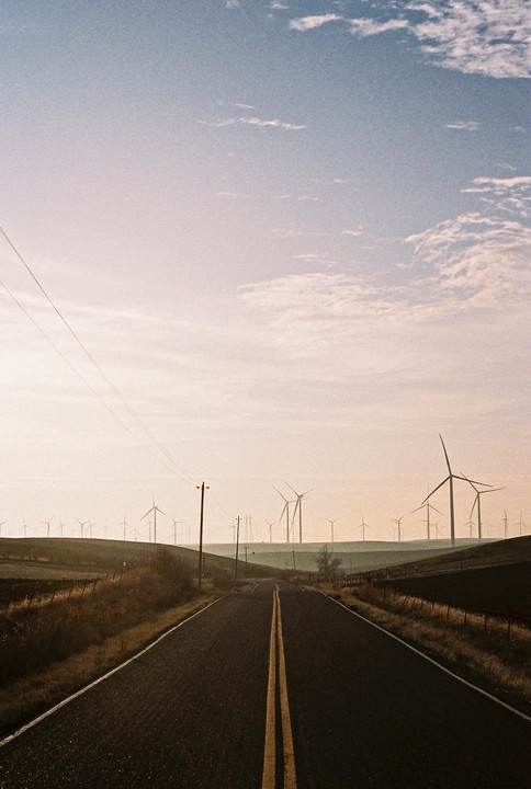 A road leads through rolling hills with numerous wind turbines under a hazy sky at sunset.