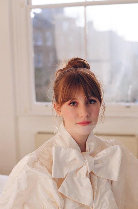 A woman with red hair and a large bow on her blouse looks directly at the viewer.