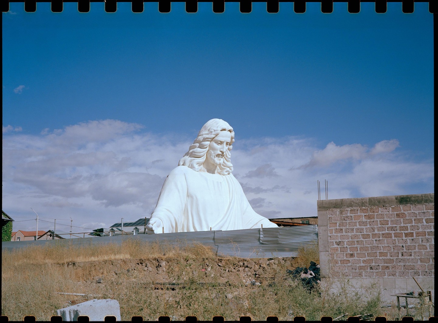 A large white statue of Jesus is visible behind a corrugated metal fence and a brick wall under a blue sky with clouds.