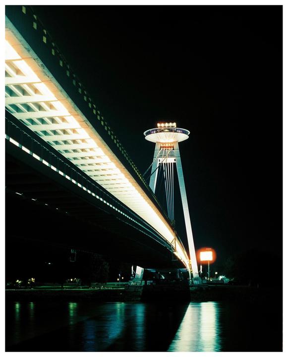 A bridge with a UFO-like structure on top is illuminated at night, reflecting in the water below.