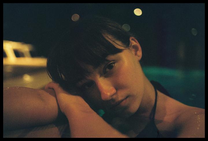 A woman with wet hair and skin rests her head on her arm in a pool at night.