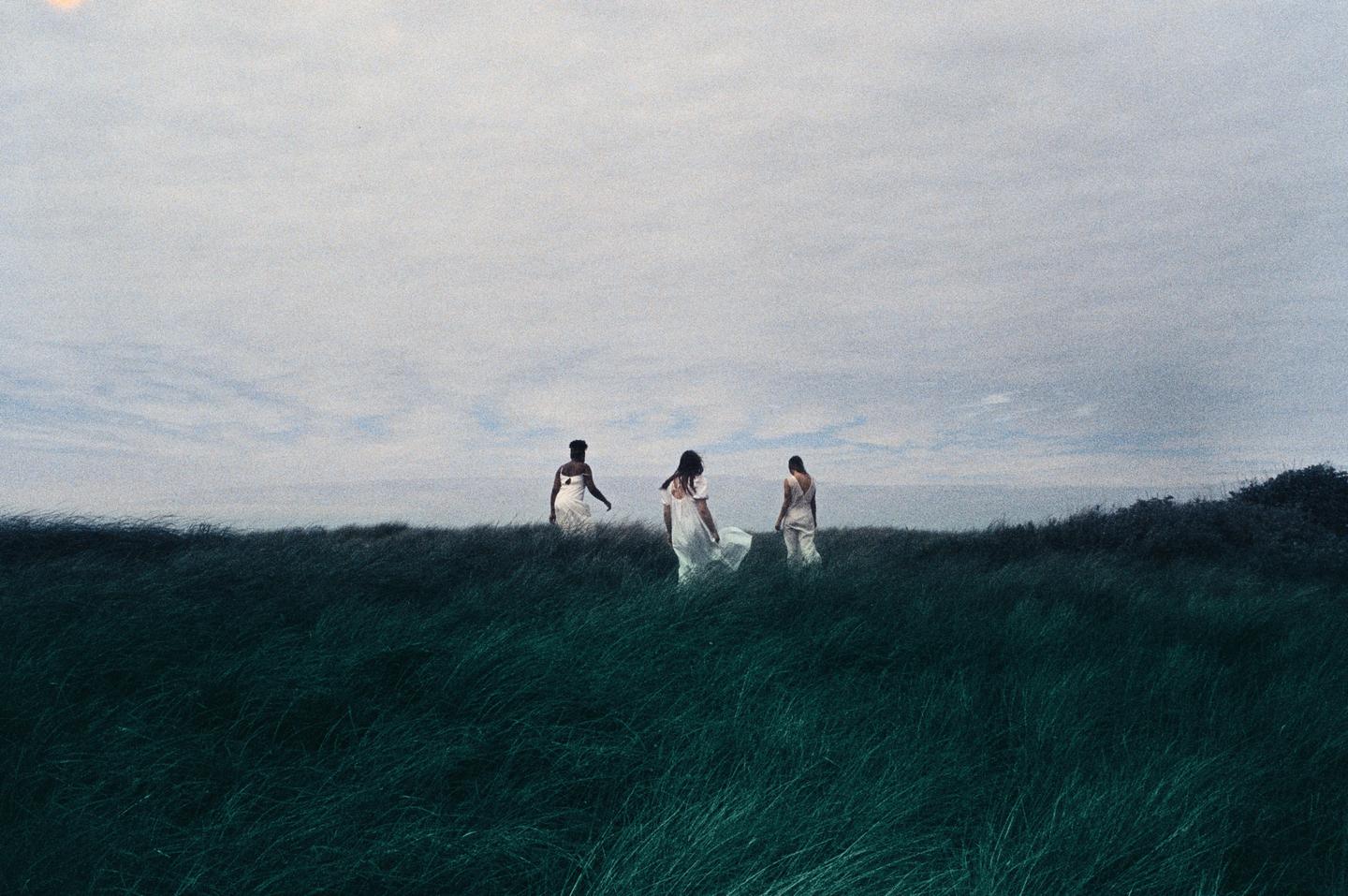 Three women in white dresses walk through a field of tall green grass under a cloudy sky.