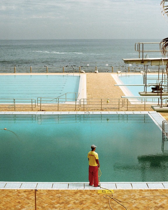 A lifeguard stands by a swimming pool overlooking the ocean with diving boards in the background.