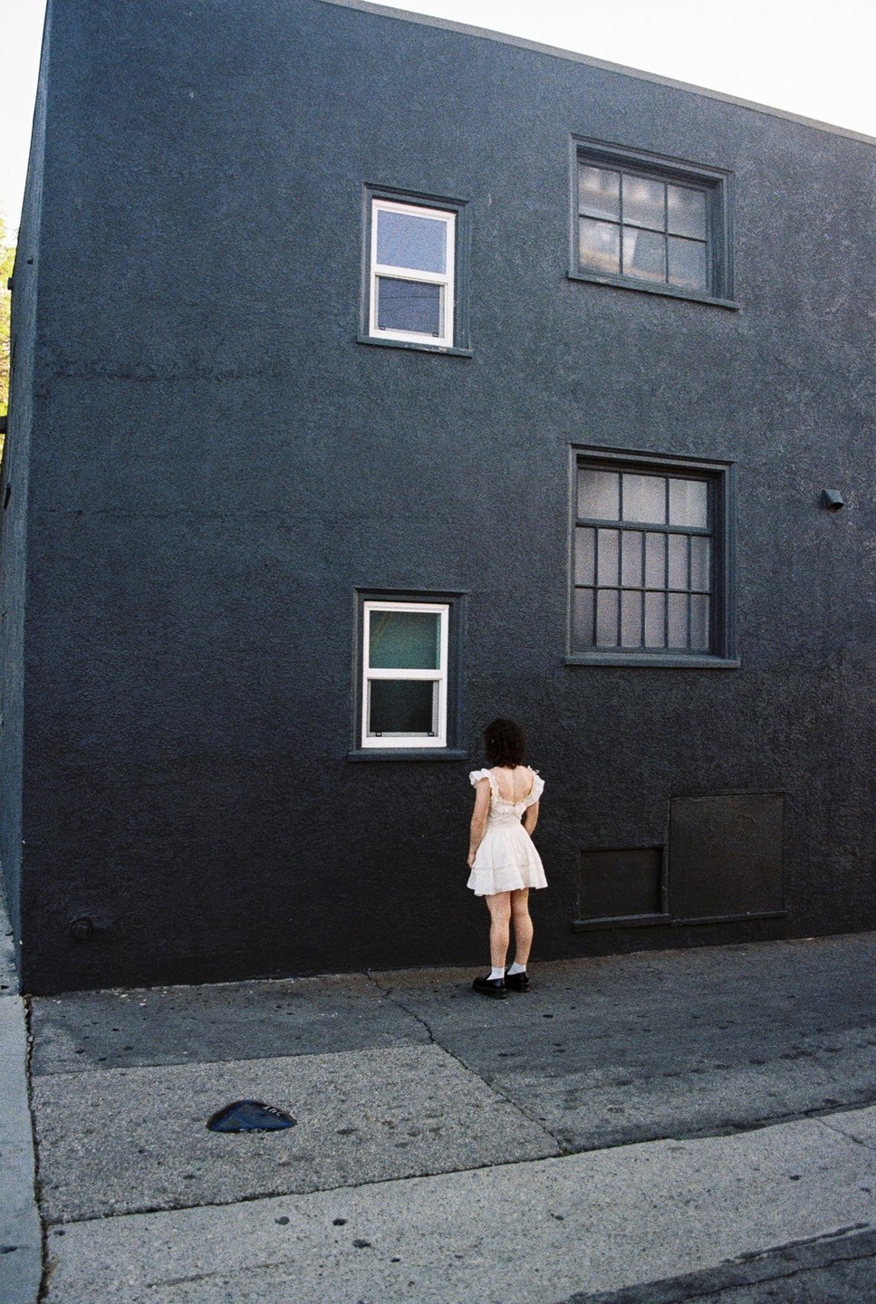 A woman in a white dress stands facing a dark building with several windows.