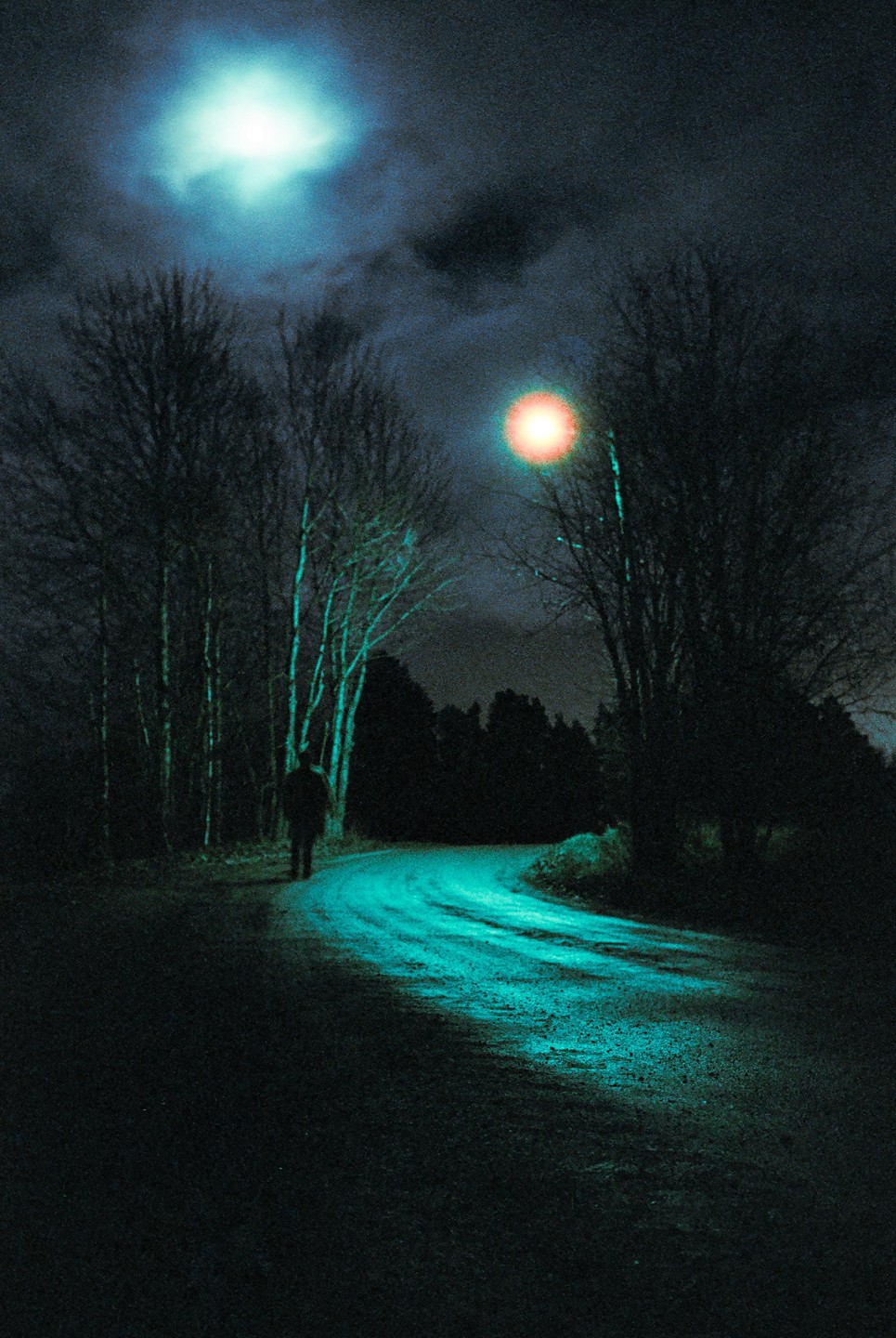 A person walks along a winding road at night, illuminated by a bright moon and a distant light source.