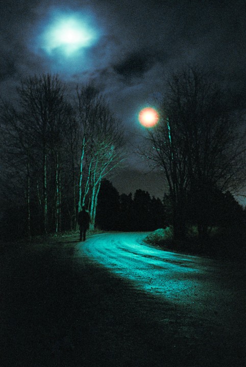 A person walks along a winding road at night, illuminated by a bright moon and a distant light source.