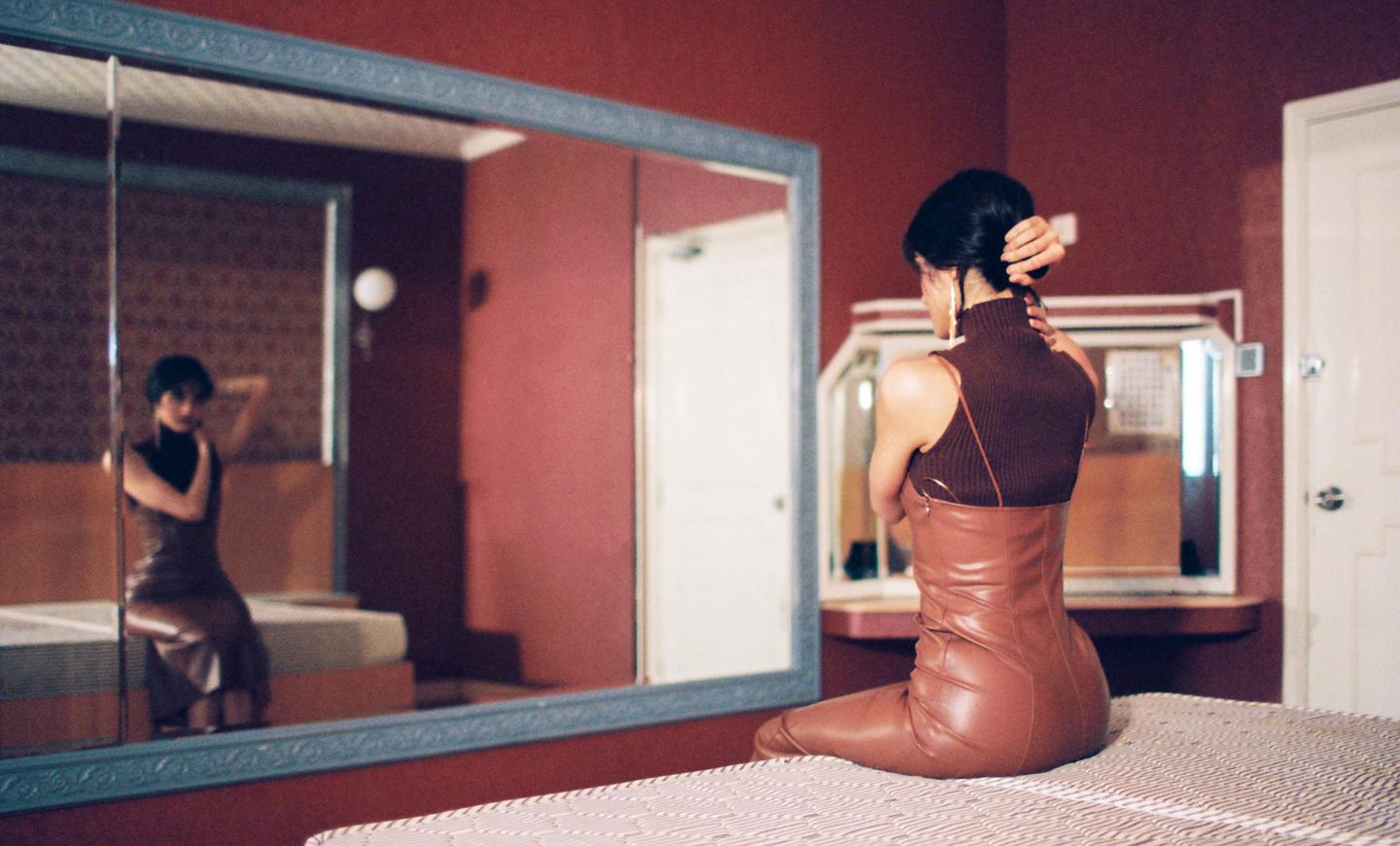 A woman in a brown dress sits on a bed, looking at her reflection in a large mirror.