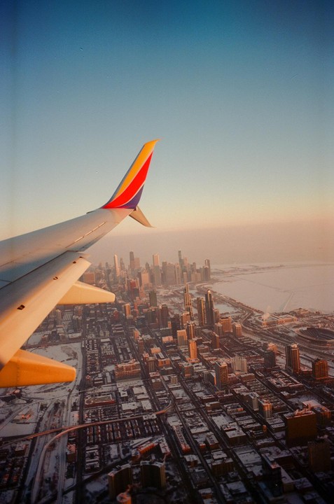 An airplane wing is visible over a snow-covered city with a lake in the background during golden hour.