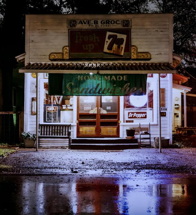A small grocery store with a "Homemade Sandwiches" sign is illuminated at night, with reflections on the wet ground in front.