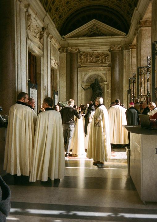 A group of men in religious robes are gathered in a grand hall with ornate architecture and a statue.