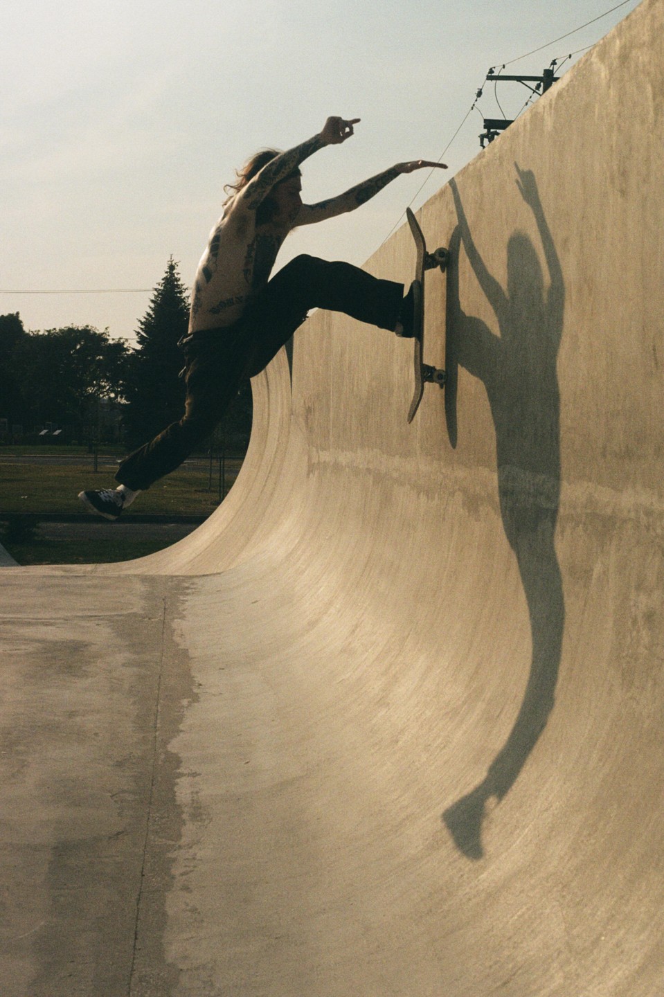 A shirtless man with tattoos is skateboarding on a concrete ramp, casting a long shadow.