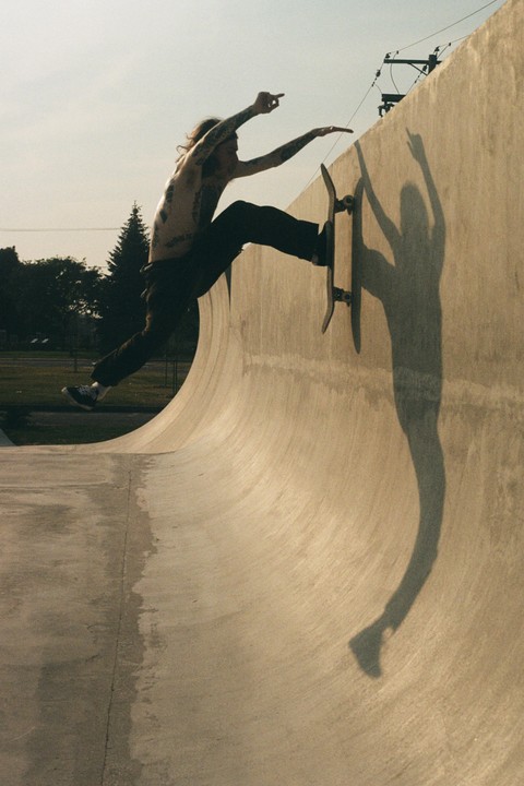 A shirtless man with tattoos is skateboarding on a concrete ramp, casting a long shadow.