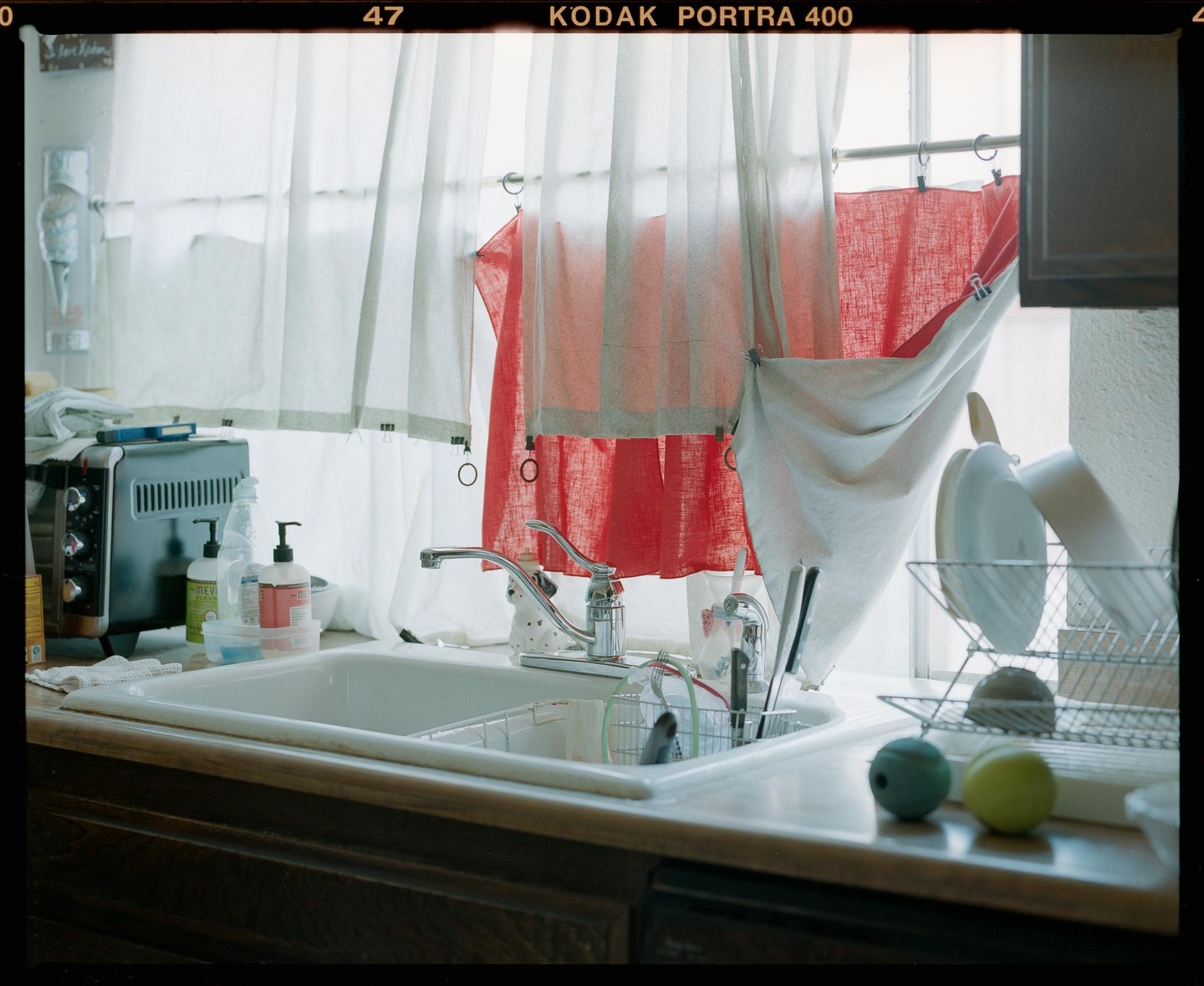 A kitchen sink is shown with a toaster oven, dish soap, and drying dishes next to a window with red and white curtains.