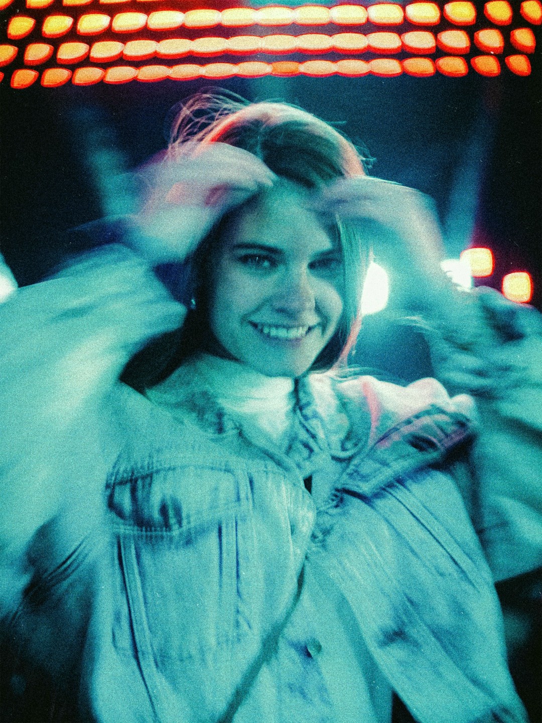 A woman with her hands in her hair smiles at the camera under orange lights.