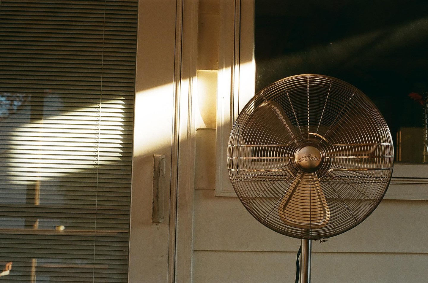 Sunlight streams through blinds and illuminates a standing fan next to a door.