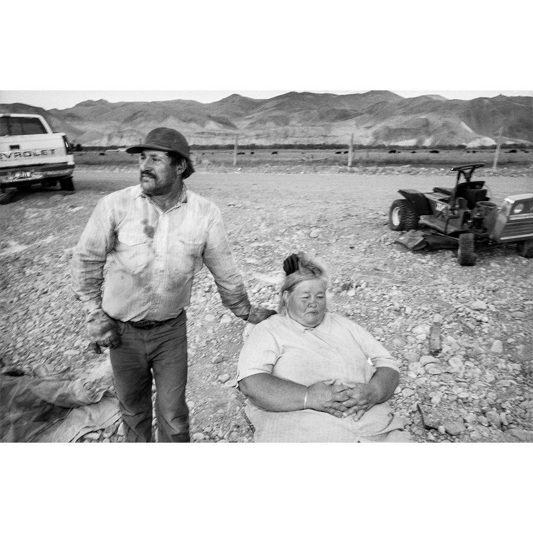 A man in a hat stands next to a seated woman on a rocky ground with mountains in the background.