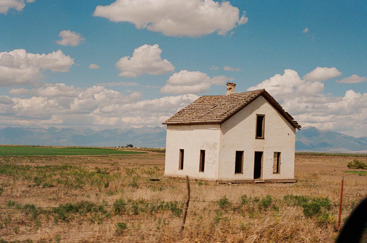 An abandoned white farmhouse stands in a dry field under a blue sky with clouds, with mountains in the distance.