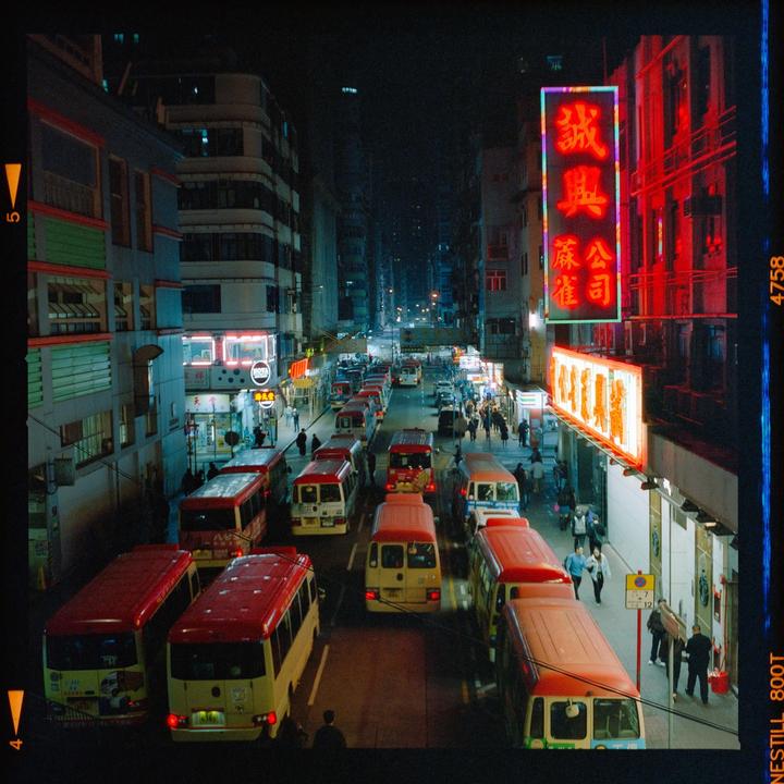 A street in Hong Kong is filled with traffic and pedestrians at night, with neon signs illuminating the buildings.