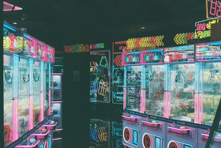 A row of claw machines with neon lights is reflected on a dark floor.
