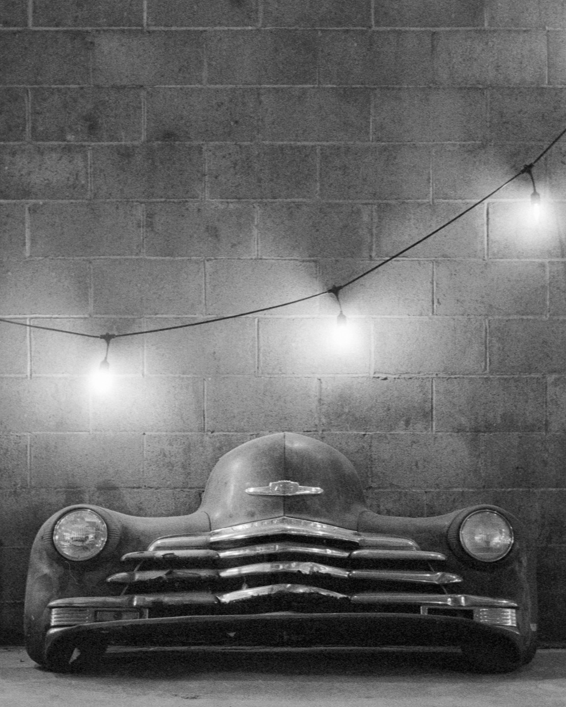 A vintage car is parked in front of a brick wall with string lights hanging above it.