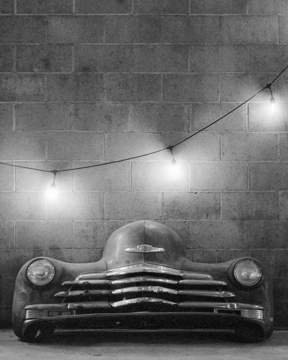 A vintage car is parked in front of a brick wall with string lights hanging above it.