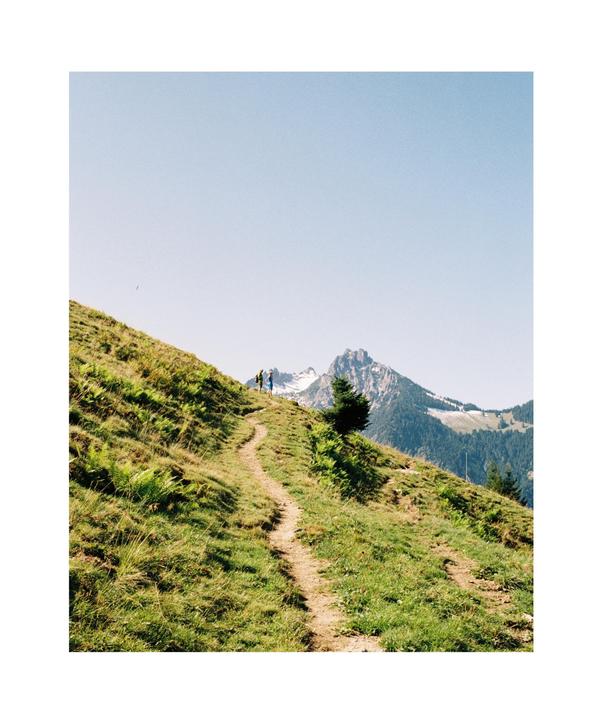 Two people walk up a winding dirt path on a grassy mountain with snow-capped peaks in the distance.