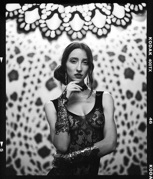 A woman in a lace top and gloves poses in front of a patterned background.