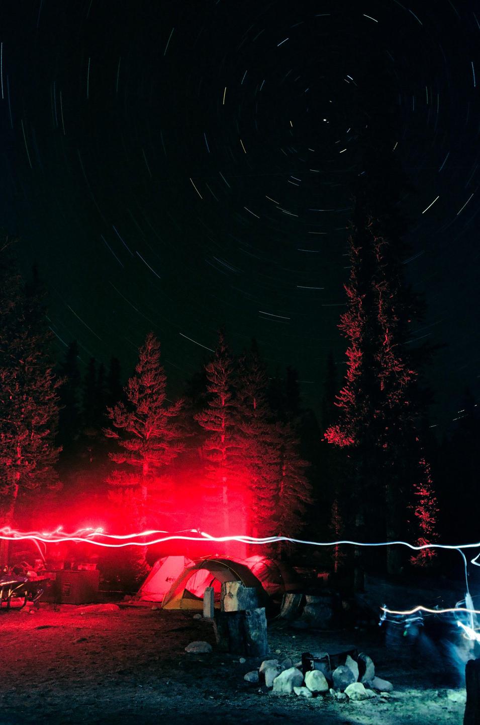 A campsite at night is illuminated by red light, with star trails visible in the dark sky above.
