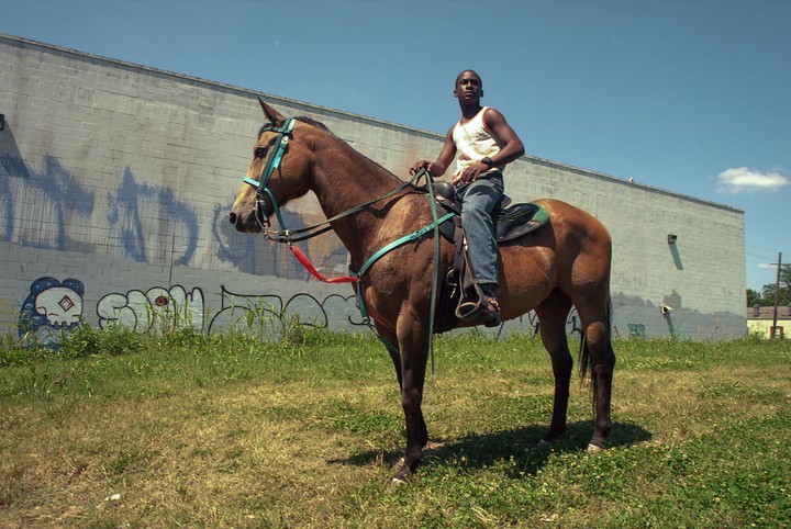 A man sits on a horse in a grassy field next to a building with graffiti.