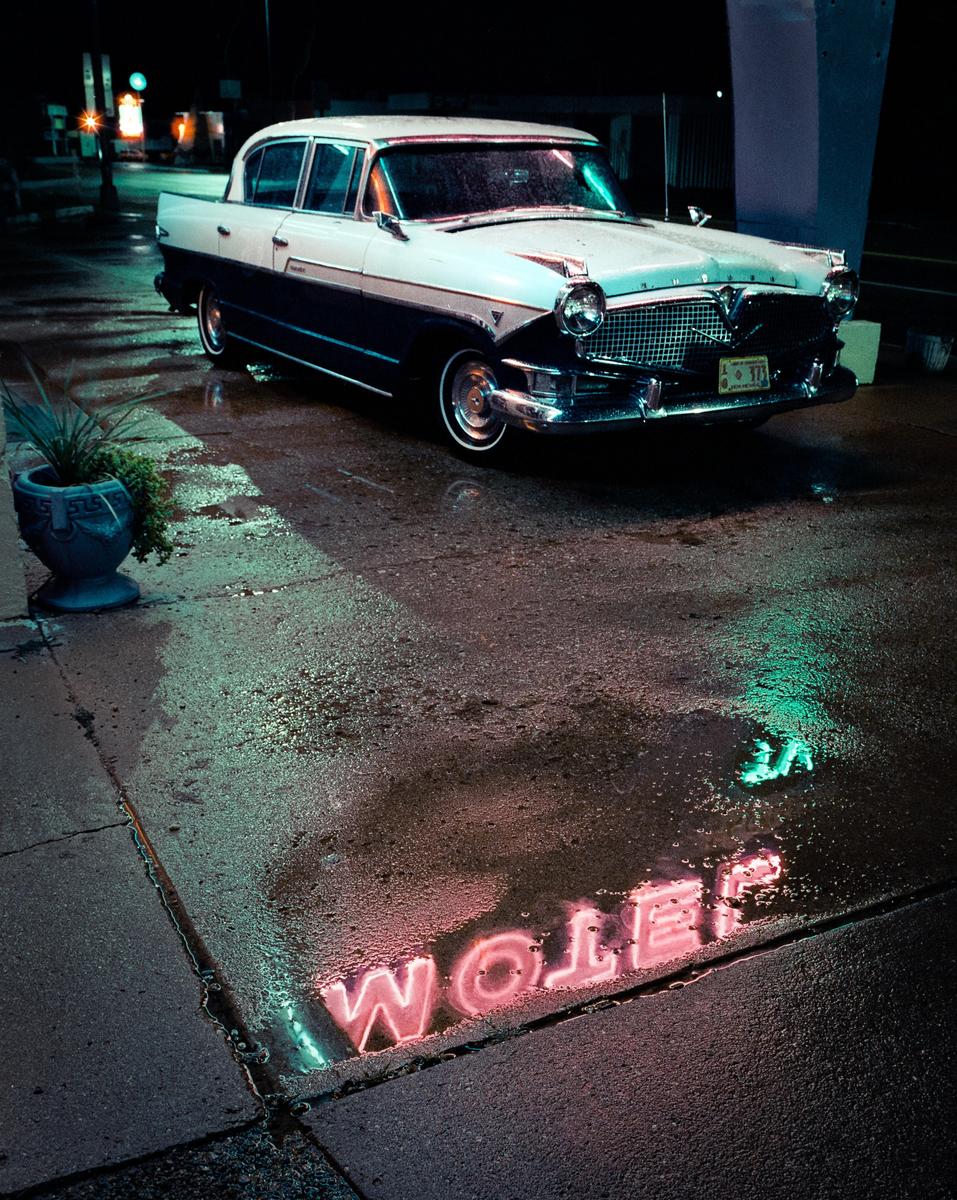A vintage car is parked on a wet street at night, with neon reflections visible in a puddle.