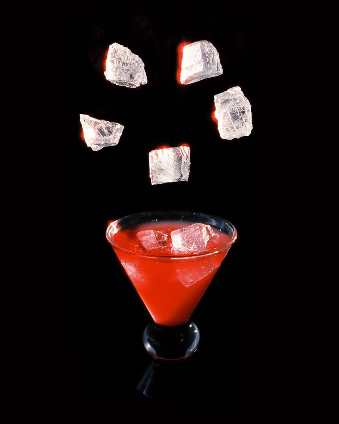 Ice cubes fall into a red cocktail in a martini glass.