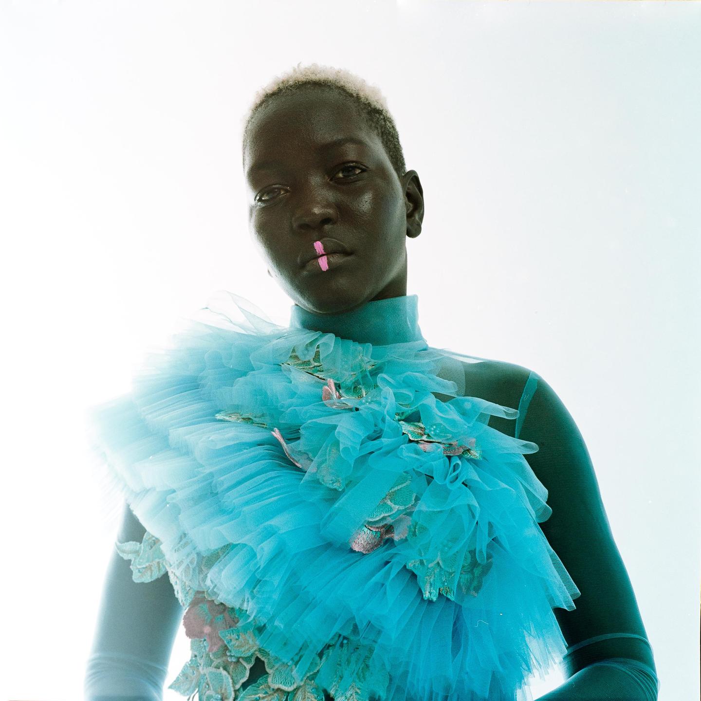 A woman with short blonde hair and a pink mark on her lip wears a blue ruffled garment.