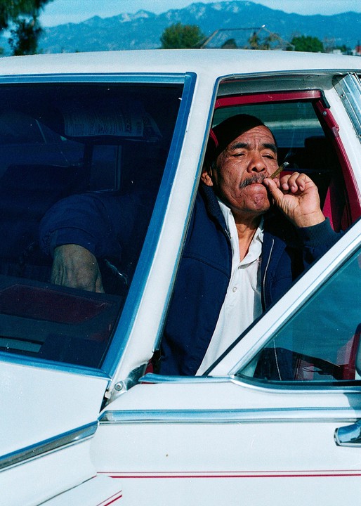 A man with a mustache sits in a white car, holding a lit cigarette to his mouth, with mountains in the background.