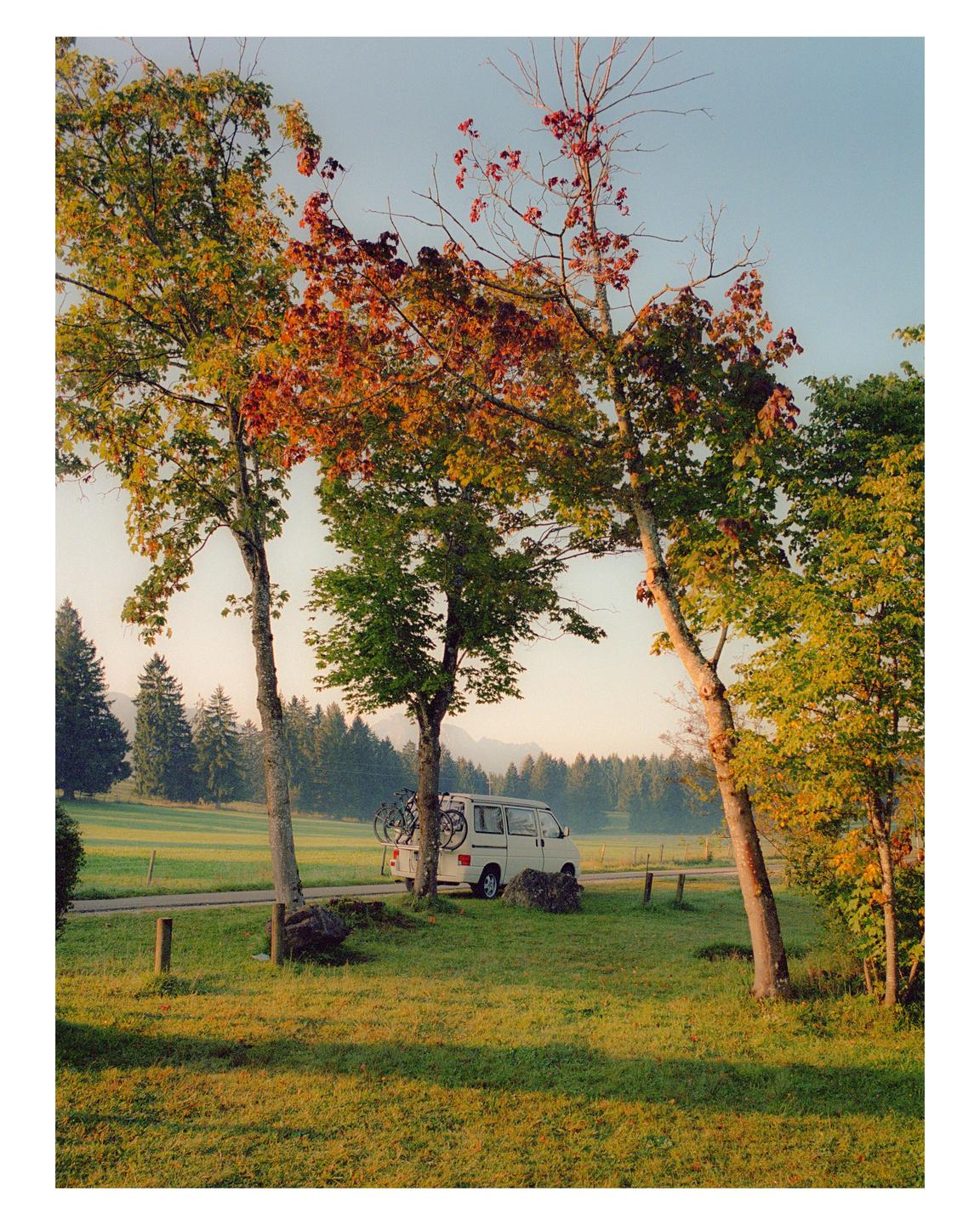 A white van with bicycles on the back is parked on a road next to a grassy field with trees showing autumn foliage.