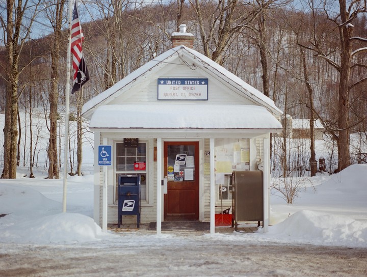 A small, snow-covered post office with an American flag flying outside stands among bare trees.