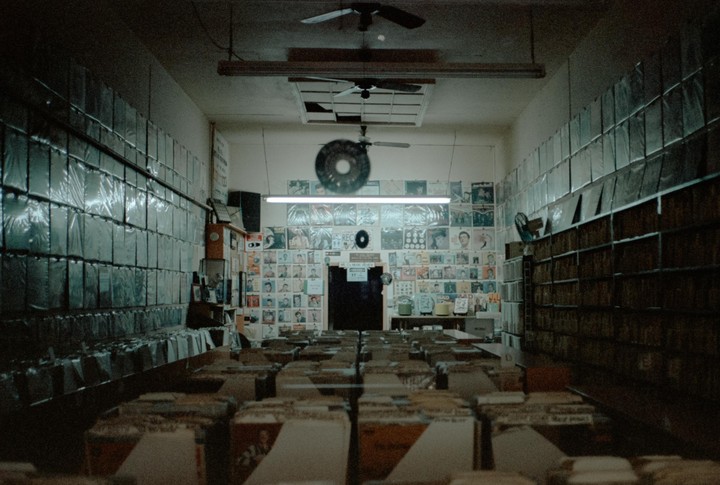 A long, narrow record store is filled with rows of vinyl records and album covers on the walls.