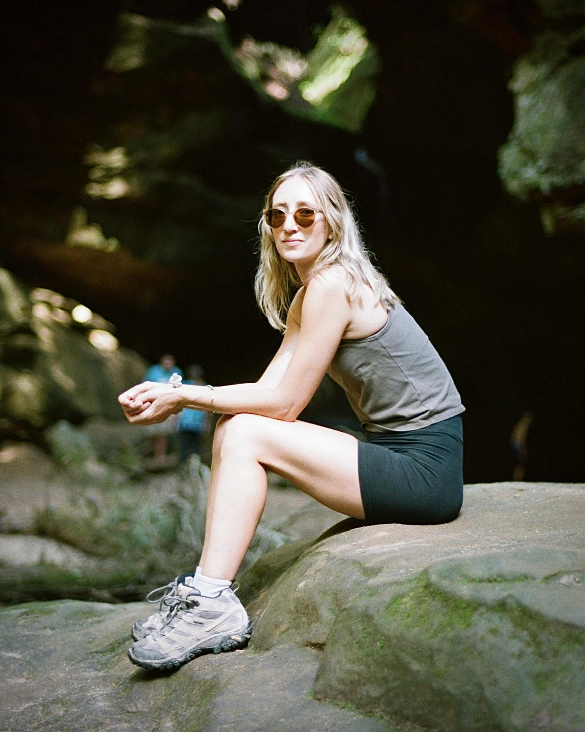 A woman wearing sunglasses, a tank top, and shorts sits on a rock in a cave.