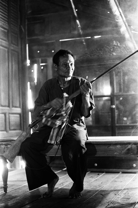 A man sits on a wooden bench, holding a long rifle and cleaning it with a cloth.