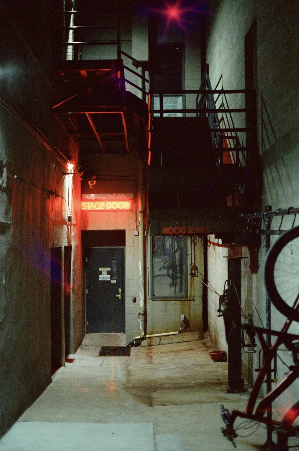 A narrow alleyway with a neon "STAGE DOOR" sign and a bicycle in the foreground is illuminated by red light at night.