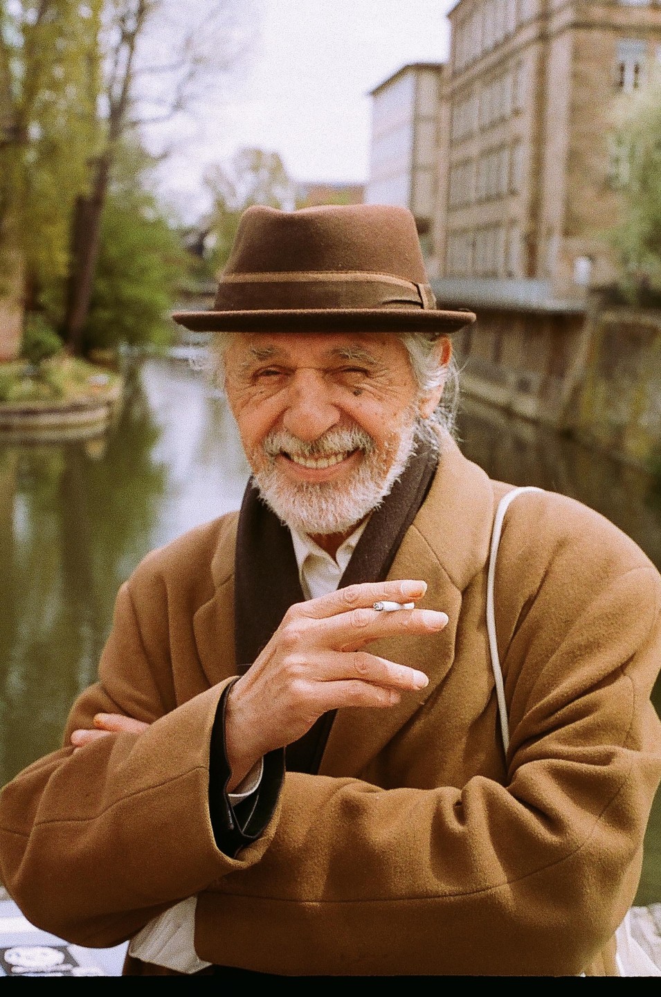 A man in a brown hat and coat smiles at the camera while holding a cigarette, with a canal and buildings in the background.