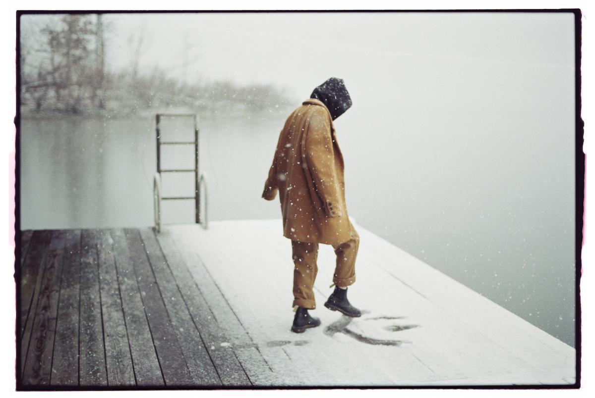 A person in a brown coat and black boots walks on a snow-covered wooden dock next to a body of water with a ladder.