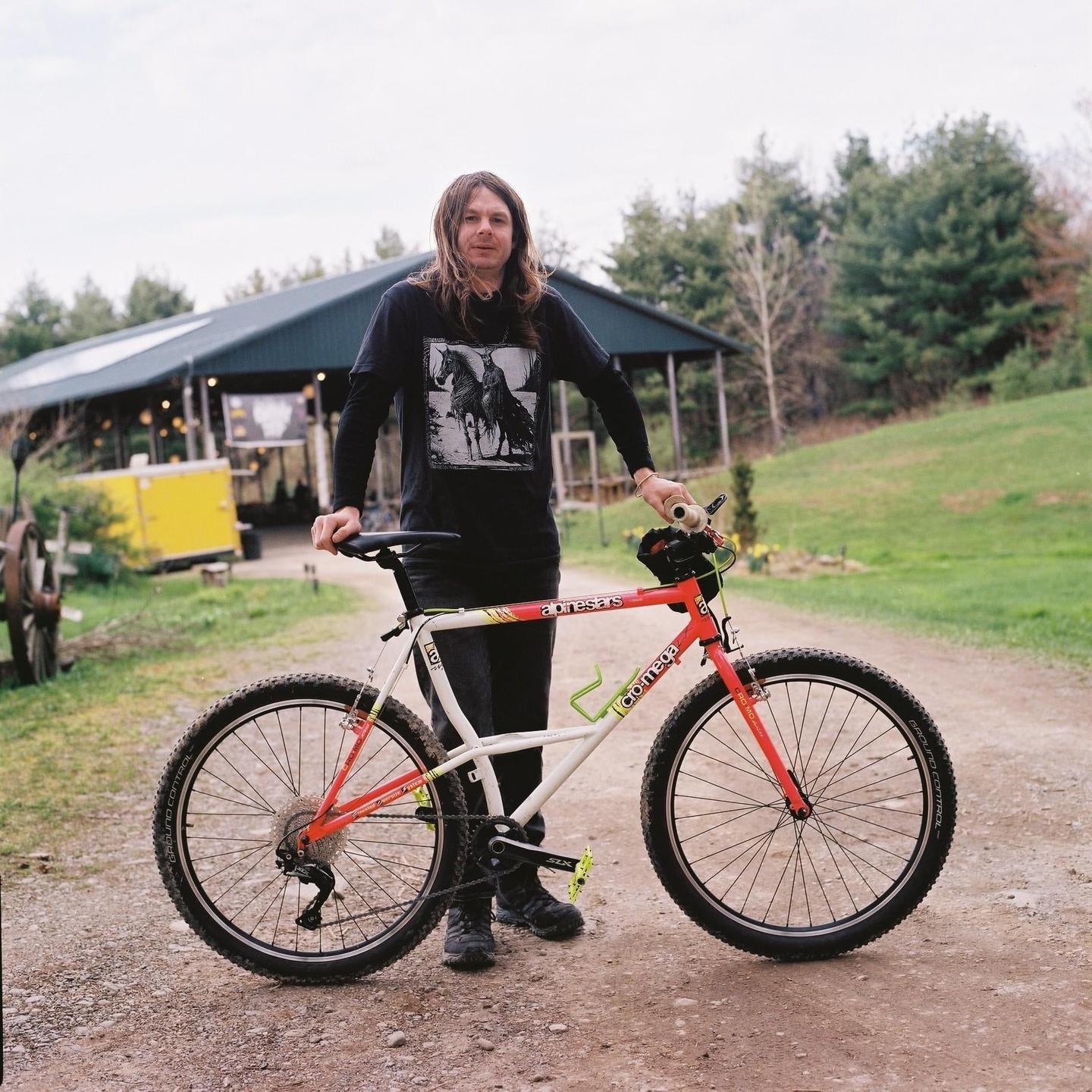 A man with long hair stands on a dirt path next to a red and white bicycle.