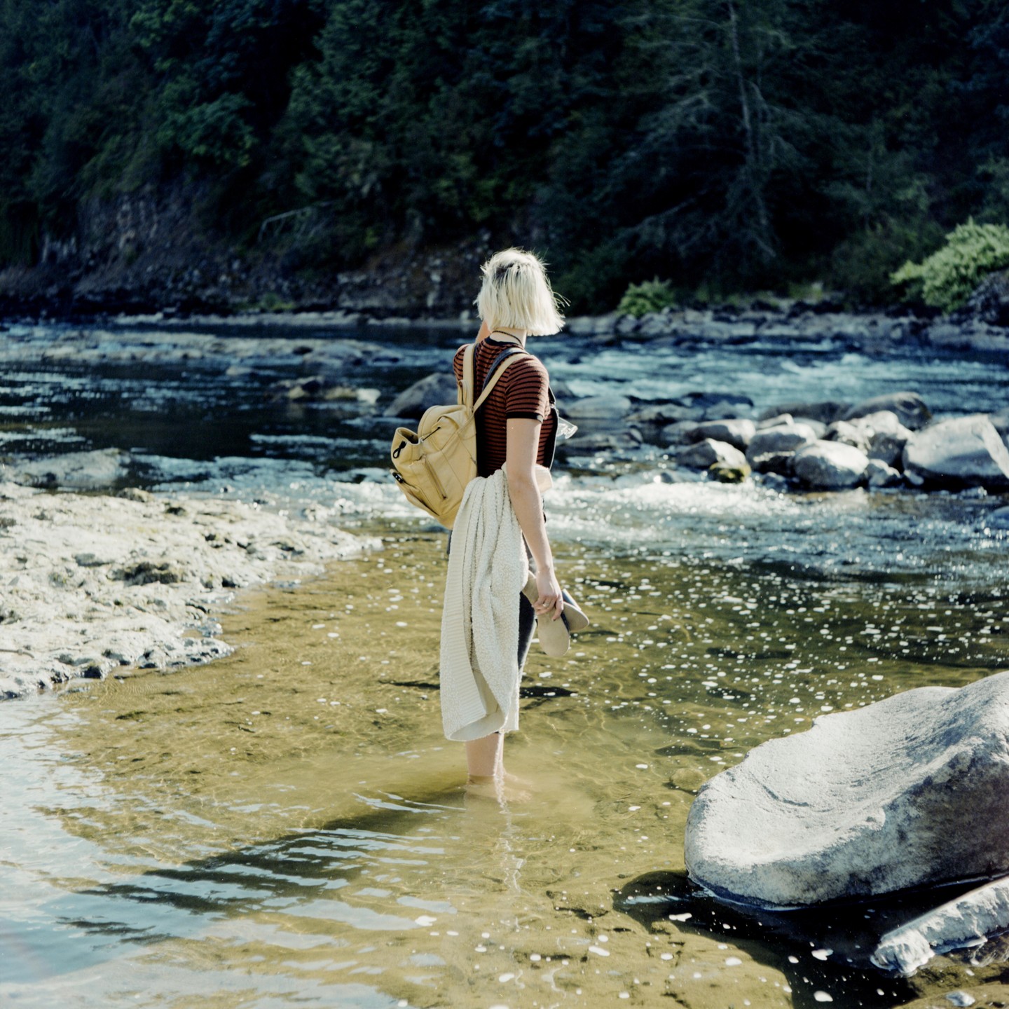 A woman stands in a shallow river with a backpack and a blanket over her arm, looking towards a forested bank.