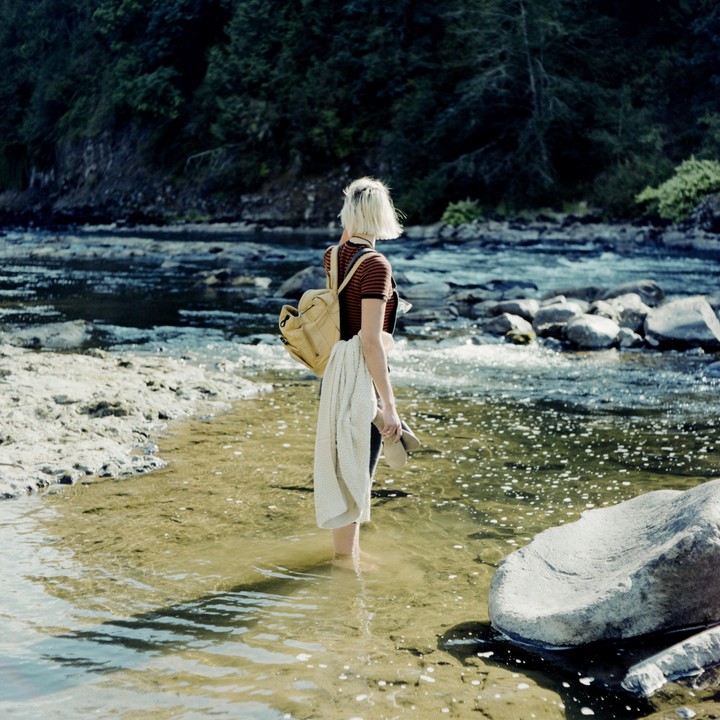 A woman stands in a shallow river with a backpack and a blanket over her arm, looking towards a forested bank.