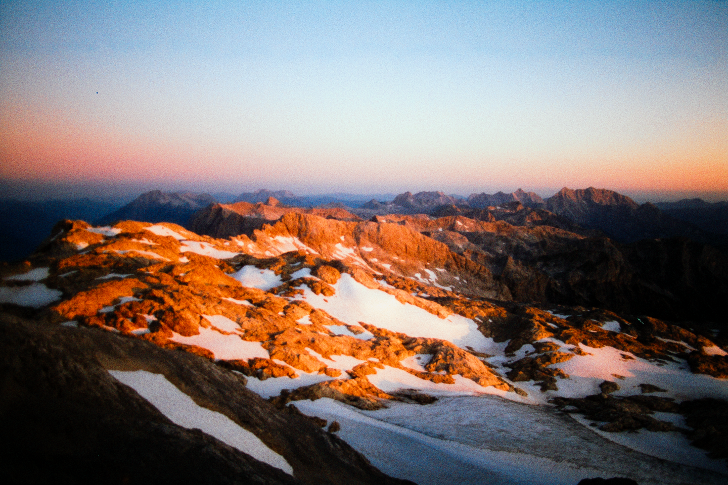A mountain range with patches of snow is illuminated by the golden light of sunset.